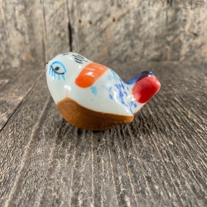 May include: A small ceramic bird figurine with blue, white, orange, and red paint. The bird has a whimsical design and a glazed finish.