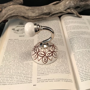 May include: A decorative white and brown floral design cabinet knob with a silver-colored metal base. The knob is on an open book with the word "MEAT" visible.