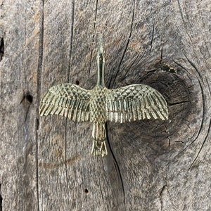 May include: A gold-colored metal crane figurine with outstretched wings. The bird is detailed with textured feathers and is set against a weathered wooden background.