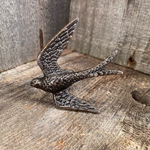 May include: Bronze-colored metal bird figurine in flight, with detailed feather textures. The bird is mounted on a wooden surface, showcasing its wings spread wide. The sculpture is a decorative item, possibly a cabinet pull or wall decor.