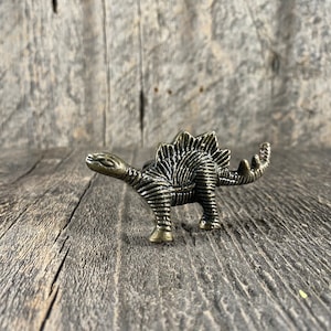 May include: A bronze dinosaur figurine with a textured surface and a spiky back. The dinosaur is standing on a wooden surface.