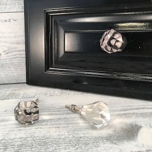Peut inclure: Gros plan sur trois boutons de meuble décoratifs. L'un est installé sur une porte d'armoire noire. Les deux autres boutons sont détachés et ont un aspect facetté en forme de cristal. Les boutons ont une quincaillerie de couleur argent.