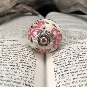 May include: A white ceramic knob with a pink and green floral design. The knob has a silver metal ring around the center.