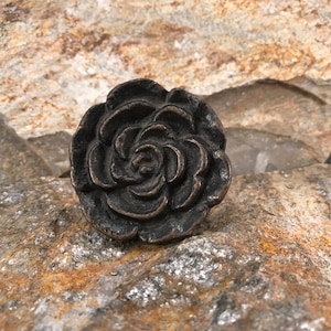 May include: A bronze-colored, rose-shaped decorative knob. The knob is highly detailed, with multiple layers of petals forming the flower. The knob is set against a textured, light brown and gray stone surface.