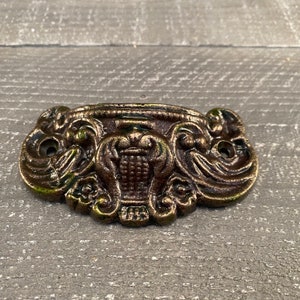 May include: A decorative cast iron drawer pull with an ornate design featuring a central floral motif and a distressed bronze finish.