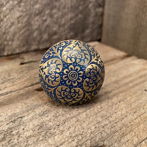 May include: A round, blue decorative object with a gold floral pattern. The object is resting on a wooden surface. The gold design is embossed on the blue surface, creating a textured appearance.