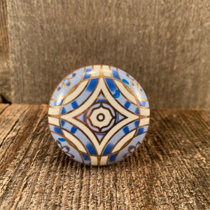 Puede incluir: Un pomo de cerámica blanca con un patrón geométrico azul y dorado. El pomo es redondo y tiene una superficie lisa.