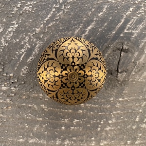 Op de afbeelding: Een ronde, zwarte knop met een gouden, ingewikkeld bloemmotief.