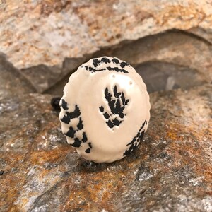 May include: A white ceramic knob with a black floral design. The knob has a scalloped edge and a screw on the back.