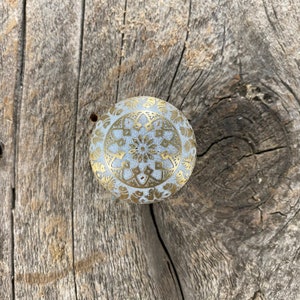 May include: A white knob with a gold mandala design. The knob is on a weathered wooden surface.