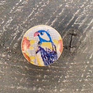 May include: A ceramic knob with a colorful bird design. The bird is blue, yellow, and purple with a red beak. The background is a light purple with a white grid pattern and pink flowers.