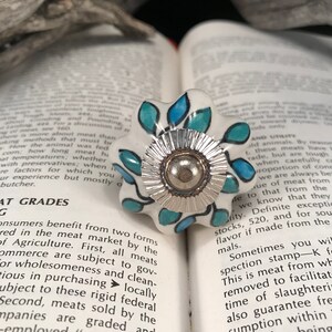 May include: A white ceramic knob with a blue and green floral design. The knob has a silver metal center with a raised, textured edge.