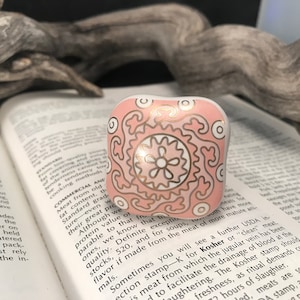 Op de afbeelding: Een roze en gouden keramische ringhouder met een decoratief bloemmotief. De ringhouder staat op een boek.