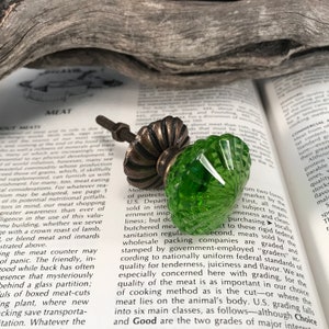 Pode incluir: Um puxador decorativo para móveis com topo de vidro esmeralda translúcido facetado e base de metal canelado em tom de bronze antigo. O hardware inclui um parafuso roscado e uma porca correspondente para uma instalação segura.