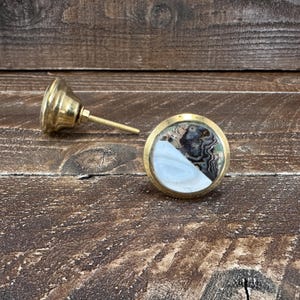 May include: A round cabinet knob with a polished brass base and a circular inlay featuring a split design. One half of the inlay is a smooth, solid white stone, while the other half displays a dark, swirling, agate-like pattern with grey and black tones.