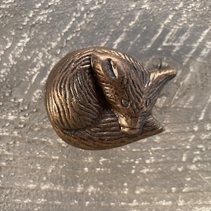 May include: Bronze-colored, decorative cabinet knob featuring a curled-up fox design. The fox is detailed with textured fur and a serene expression. This unique hardware adds a touch of nature-inspired charm to furniture.