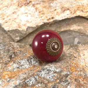 Może przedstawiać: Ceramiczny gałka w kolorze ciemnej czerwieni z akcentem mosiężnym. Gałka ma dekoracyjny, okrągły wzór z wypukłym środkiem.