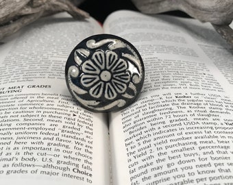 Könnte beinhalten: Ein schwarzer und weißer Keramik-Knopf mit einem floralen Design. Der Knopf ist rund und hat ein erhabenes Design. Der Knopf liegt auf einem Buch.