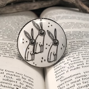 May include: A white ceramic knob with a black and white illustration of three rabbits. The rabbits are drawn in a simple, stylized manner.