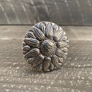 May include: A silver metal knob with a detailed sunflower design. The knob is round and has a raised center.
