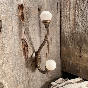 May include: A double wall hook featuring a textured, dark bronze-toned metal frame with two curved arms. Each arm terminates in a smooth, spherical white knob. The base of the hook is circular and secured with two visible black screws.