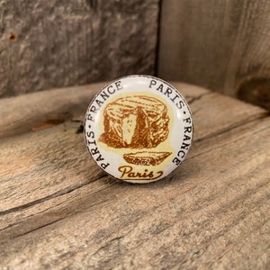 May include: A white and cream-colored round cabinet knob with a brown illustration of cheese and the word "Paris" in a vintage style. The text "Paris France" is printed around the edge of the knob.