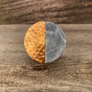 Peut inclure: Un bouton de meuble circulaire au design divisé. Une moitié est en métal doré martelé avec une surface texturée, tandis que l'autre moitié est composée d'un matériau gris foncé lisse ressemblant à de la pierre avec une finition mouchetée.