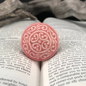 May include: A pink ceramic ring with a white design. The design is a repeating pattern of swirls and dots.