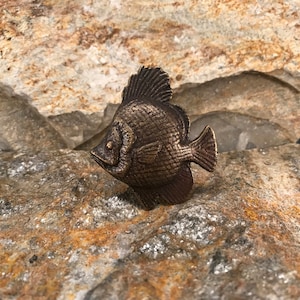 May include: A bronze-colored fish figurine with a detailed, textured surface. The fish has a large, rounded fin and a small, pointed tail.