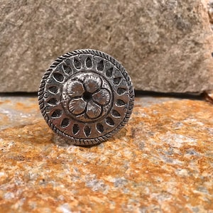 May include: A silver ring with a floral design. The ring is round and has a raised flower in the center. The flower has five petals and a small hole in the center. The ring is surrounded by a border of small, teardrop-shaped designs.
