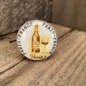May include: White ceramic knob with a brown and yellow illustration of a wine bottle and glass. The knob is decorated with the text "Paris. France. Paris. France" in a black circular border.