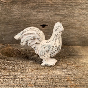 May include: A white cast iron rooster figurine with a distressed finish. The rooster has a detailed tail, body, and head. The figurine is a decorative item, likely for a farmhouse or rustic style home decor.