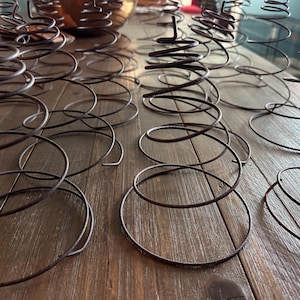 May include: Close-up of several rusty metal springs arranged on a wooden surface. The springs are dark brown and coiled, with a spiral design. The image shows a collection of vintage or antique hardware components.