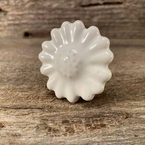 Pu&ograve; includere: Pomello per armadio a forma di margherita in ceramica bianca. Il pomello ha una superficie dettagliata e strutturata con un'area centrale circolare leggermente rialzata. I petali della margherita sono smerlati.
