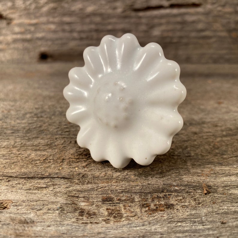Pu&ograve; includere: Pomello per armadio a forma di margherita in ceramica bianca. Il pomello ha una superficie dettagliata e strutturata con un'area centrale circolare leggermente rialzata. I petali della margherita sono smerlati.
