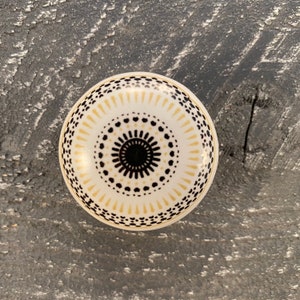 Op de afbeelding: Een witte keramische knop met een geometrisch patroon in zwart en goud. De knop is rond en heeft een reliëfdesign.
