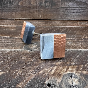 May include: Two square cabinet knobs featuring a split design with one half made of smooth grey marble and the other half made of hammered copper metal. Each knob includes a threaded metal screw extending from the back for installation purposes.