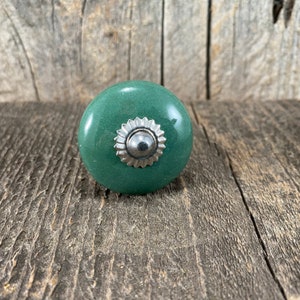 May include: A green ceramic knob with a silver decorative accent. The knob is round and has a smooth surface.