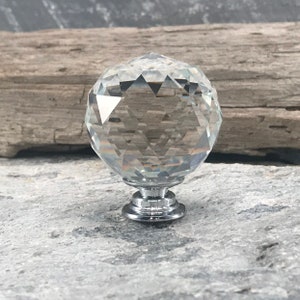 May include: A clear, faceted crystal knob with a chrome base. The knob is spherical with multiple flat surfaces, reflecting light. The chrome base has a tiered design. The knob is likely for furniture or decorative use.