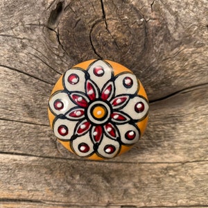 May include: A round ceramic knob with a yellow background and a white flower design. The flower has red petals and a yellow centre with black accents.