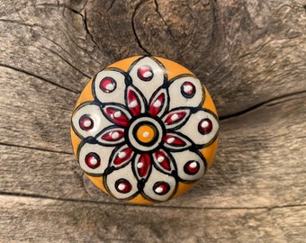 Op de afbeelding: Een ronde keramische knop met een gele achtergrond en een wit bloemenontwerp. De bloem heeft rode bloemblaadjes en een gele kern met zwarte accenten.