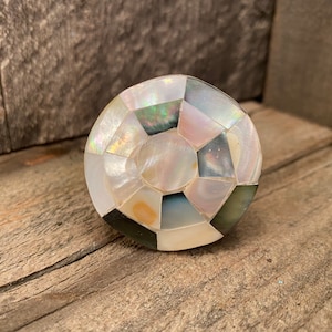 May include: A round, decorative cabinet knob with a mosaic design. The knob is made of iridescent mother-of-pearl tiles in shades of white, black, and gray. The knob is set against a rustic wooden surface.