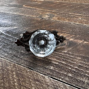 Peut inclure: Une poignée de tiroir composée d'un bouton en verre transparent à facettes avec un design géométrique circulaire. Le bouton est fixé sur une plaque arrière en métal brun foncé ornée de motifs complexes en forme de feuilles.
