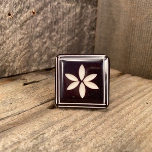 Puede incluir: Una caja cuadrada de color púrpura oscuro con un diseño floral blanco en el centro. La caja tiene un borde blanco y está colocada sobre un fondo de madera envejecida. El diseño floral es una flor de seis pétalos.