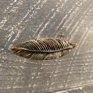 May include: A gold-toned metal feather-shaped drawer pull. The pull has a detailed design and is perfect for adding a touch of rustic charm to any home decor.