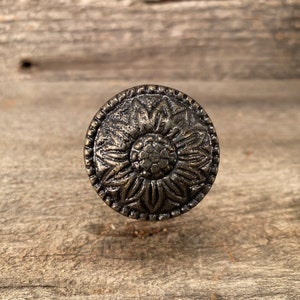 Może przedstawiać: Okrągła gałka do szafki w stylu antycznym z kwiatowym wzorem. Gałka wykonana jest z metalu z ciemnobrązowym wykończeniem. Kwiatowy wzór jest wytłoczony, dodając tekstury i szczegółów. Gałka znajduje się na drewnianej powierzchni.