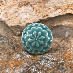 Puede incluir: Un pomo de cerámica azul turquesa con un diseño floral blanco. El pomo tiene una superficie texturizada en relieve y un borde blanco.