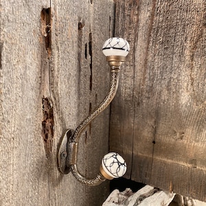 May include: A decorative wall hook with a brass finish. The hook features two white ceramic knobs with black abstract designs. The hook is mounted on a weathered wooden surface, adding a rustic touch to the design.