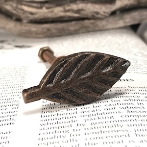 May include: A bronze leaf-shaped cabinet knob with a threaded screw. The knob has a rustic, aged finish.