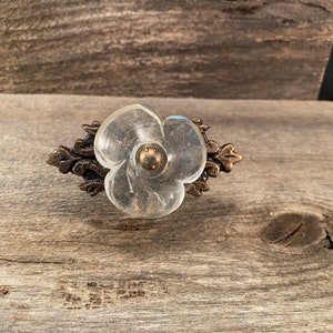 Pode incluir: Um puxador de armário transparente em forma de flor, com um centro de cor latão e um suporte decorativo de metal cor bronze. O puxador está sobre uma superfície de madeira cinza envelhecida.
